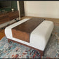 An oversized wood ottoman table with a dark walnut stained tray on top of an ottoman, placed in a room with a wooden floor and a multicolored area rug.